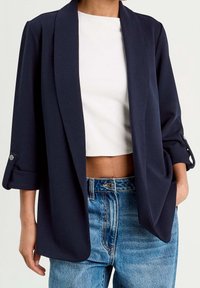 Blazer bleu marine à coupe décontractée, confectionné dans un tissu doux. Il présente un col châle, des manches raccourcies et une coupe ajustée. Porté avec un jean taille haute.