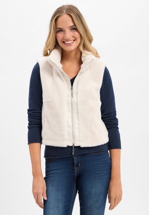 Faux fur vest in crèmekleur met hoge kraag en volledige rits aan de voorkant. Het model draagt een marineblauw topje eronder, gestyled met strakke spijkerbroeken.