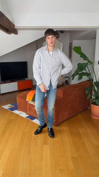 Mladenič v črtasti majici in kavbojkah stoji v dnevni sobi z oranžnim kavčem, veliko rastlino, televizijo in lesenim podom.