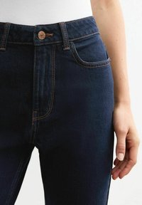 Jeans en denim bleu foncé avec des surpiqûres orange contrastantes, taille haute, poches avant et fermeture à bouton en cuivre. Texture lisse.