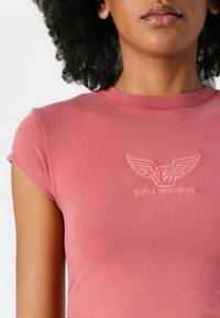 Rosa, tettskjorte med korte ermer og rund hals, med et brodert logo med vinger i lys rosa.