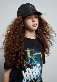 Căciulă neagră cu un logo alb, purtată cu un tricou negru care are un print grafic colorat și părul buclat care încadrează fața.