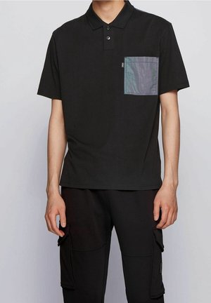 Schwarzes Polohemd aus Baumwolle mit flachem Kragen, zwei Knöpfen und einer grauen, irisierenden Tasche. Kombiniert mit schwarzer Cargohose.