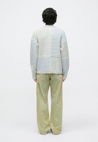 Világosszürke és kék színű kötött pulóver texturált mintákkal; oldalán cipzárokkal díszített, széles szárú, világoszöld farmernadrággal párosítva.