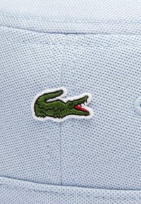 Tejido texturizado azul claro con un logotipo de cocodrilo bordado en verde con acentos rojos, contorneado en blanco. El diseño incluye costuras visibles.