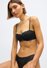 Čierny súbor spodnej bielizne z čipky zahŕňajúci ramienkovú podprsenku s kvetinovými vzormi a zodpovedajúce nohavičky. Materiály zahŕňajú priehľadnú čipku a jemnú tkaninu.