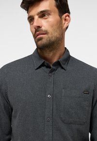 Graues Hemd mit Knopfverschluss aus strukturiertem Stoff, ausgestattet mit einer aufgesetzten Brusttasche, Kragen und einem kleinen Markenetikett auf der Vorderseite.