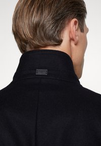 Homme aux cheveux châtains clairs tourné de dos, portant un manteau noir à col montant avec une petite étiquette rectangulaire sur le col arrière.