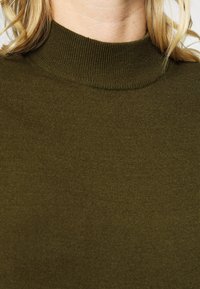 Olivgrön, ribbad polotröja. Mjuk material med en åtsittande design och hög krage. Näroptagen bild visar textur och sömdetaljer.