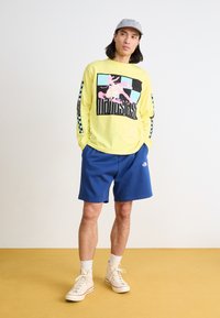 Chemise jaune à manches longues avec imprimé graphique, accents noirs et bleus. Un short bleu et des baskets beiges complètent la tenue.