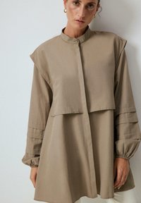 Khaki koszula zapinana na guziki z kołnierzykiem, z warstwowymi rękawami i luźnym krojem. Wykonana z lekkiego materiału o gładkiej teksturze.