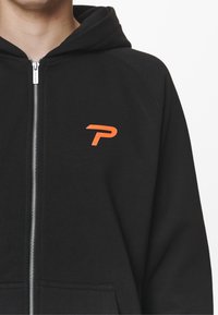 Felpa con zip nera realizzata in morbido misto cotone, con un logo arancione a forma di 'P' sul lato sinistro del petto e una tasca frontale.