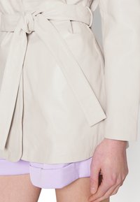 Trench-coat en cuir gris clair avec une ceinture à nouer. Le manteau a une texture lisse et un design classique, porté par-dessus un short lavande.