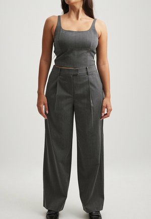 Kvinde iført en mørkegrå ærmeløs crop top og højttaljede, vidtfavnende bukser, stående mod en ensfarvet baggrund.