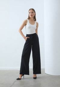 Black wide-leg pants with an elastic waistband and drawstring, paired with a white tank top and black high-heel shoes on a smooth floor.