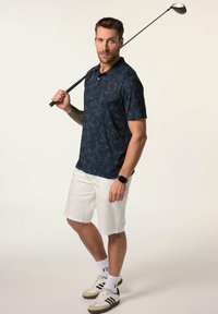 Námornícká polo košile s jemným vzorem, kombinovaná s bílými šortkami. Model drží golfovou hůl; má na sobě tenisky a hodinky, s viditelnými ponožkami.