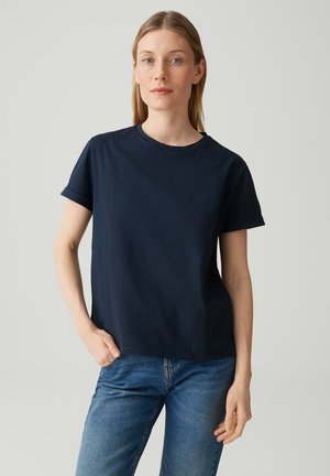 Vrouw met stijl blond haar draagt een marineblauw T-shirt met korte mouwen en een blauwe spijkerbroek, staand met één hand in haar zak tegen een effen achtergrond.