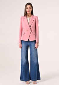 Rosa zweireihiger Blazer mit goldenen Knöpfen, zwei Fronttaschen, über einem hellen Oberteil getragen und kombiniert mit weit geschnittenen blauen Denim-Jeans.