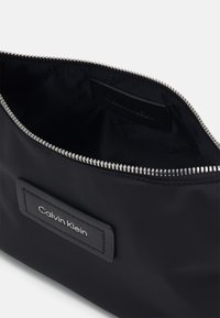 Musta kangaskosmetiikkapussi, jossa on hopeavetoketju. Sillä on pehmeä rakenne ja suorakulmainen muoto, ja sisällä on musta Calvin Klein -merkki.