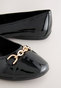 Black patent leather women's flats with gold-tone chain detail on rounded toe, smooth sole, and low heel.