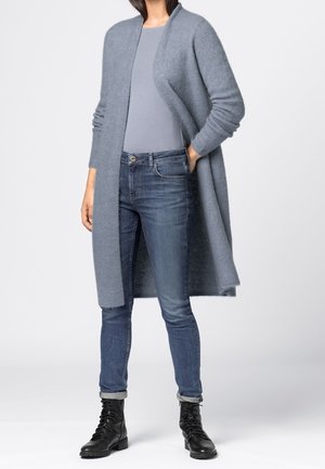 Person iført en lang grå cardigan med åben front, lysegrå top, tætsiddende blå jeans og sorte snørebåndsstøvler, står med den ene hånd i lommen.
