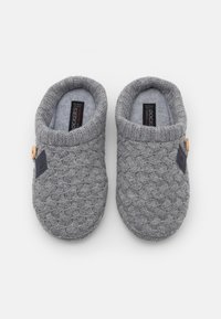 Chaussons tricotés gris avec un motif tressé, texture douce, forme arrondie ouverte, étiquette noire contrastante et détail de bouton en bois.