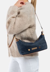 Sac à épaule en denim bleu marine avec une bandoulière en cuir, quincaillerie dorée, fermeture éclair et deux accents hexagonaux à l'avant. Texture douce.