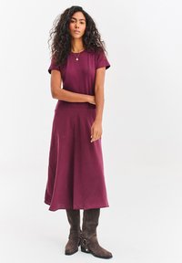 Bordowa sukienka z krótkimi rękawami, rozkloszowanym dołem w kształcie litery A i gładką fakturą. Do tego brązowe kozaki za kolano z klamrami.