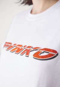 Bílá bavlněná tričko s nápisem "PINK" napsaným tučným černým a oranžovým písmem, ozdobené červenými kamínky. Krátký rukáv.