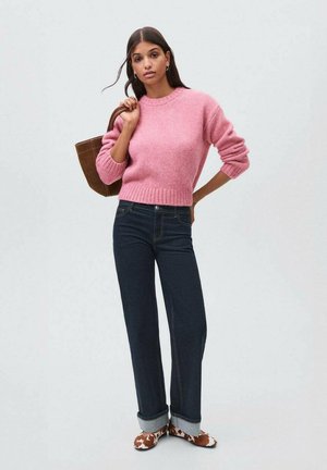 Maglione cropped rosa con orlo a costine, jeans blu scuro con risvolti e calzature con fantasia. La modella tiene una borsa tote marrone.