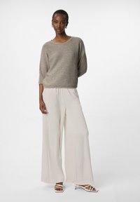 Maglione in maglia marrone chiaro con maniche corte, caratterizzato da sottili dettagli scintillanti. Abbinato a pantaloni a gamba larga di colore crema e tacchi bianchi con cinturini.
