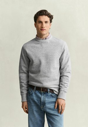 Junger Mann mit kurzen braunen Haaren, der einen hellgrauen Sweatshirt über einem gestreiften Hemd und blaue Jeans trägt, steht vor einem einfarbigen beigen Hintergrund.
