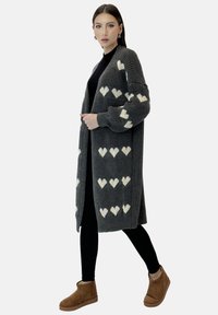 Cardigan in maglia grigia con motivi a cuore bianchi, davanti aperto, texture a coste, maniche lunghe. Leggings neri e stivaletti marroni completano l'outfit.