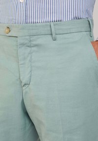 Pantaloni di un blu chiaro in tessuto liscio, con chiusura a bottone, parte frontale liscia e tasche laterali, abbinati a una camicia a righe.
