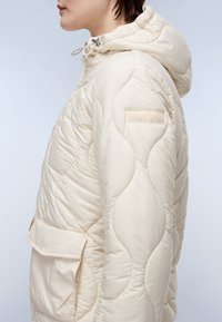 Giacca trapuntata beige con cappuccio, dotata di una tasca frontale, superficie testurizzata e piccolo logo sulla manica. Materiale leggero.