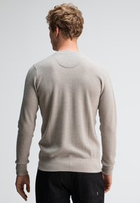 Man met krullend lichtbruin haar die een gestructureerde beige trui en zwarte broek draagt, met zijn gezicht weg van een eentonige lichte achtergrond.