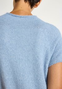 Henkilö, joka on pukeutunut lyhythihaiseen, vaaleansiniseen pörröiseen neuleeseen, joka paljastaa yläselän ja kaula-aukon, jossa on pieni sullus kaulassa.