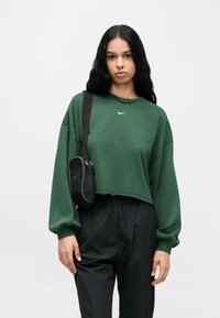 Πράσινο cropped φούτερ με μακριά μανίκια και λευκό λογότυπο Nike, συνδυασμένο με χαλαρά μαύρα παντελόνια και μικρή μαύρη τσάντα χιαστί.