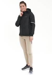 Sort zip-jakke med hætte og reflekterende detaljer, parret med beige bukser og sorte sneakers. Glat tekstur, atletisk pasform.