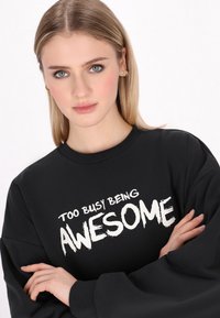 Fekete pulóver, fehér felirattal: "TÚL FOGYOSZTOTTAN CSODÁLATOS." Hosszú ujjú és kerek nyakú. Egyszerű design, sima textúra.