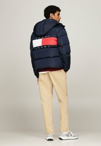 Giubbotto puffer blu navy con una striscia rossa e bianca e il testo "TOMMY JEANS" sulla schiena, abbinato a pantaloni beige chiaro e sneakers grigie.
