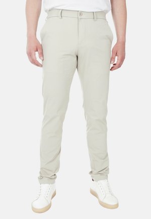 Pantaloni beige chiaro in una vestibilità slim con fronte piatto, passanti per cintura e chiusura a bottone, abbinati a sneakers bianche. Tessuto dalla consistenza liscia.
