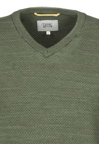 Pull en maille vert avec un col en V, affichant un motif texturé et une étiquette jaune sur le col. L'étiquette indique "camel active", taille L.