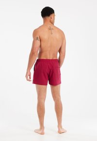 Bordeauxkleurige zwembroek met een elastische tailleband, zijzakken en een achterzak. Gladde textuur met minimale branding op het onderbeen.