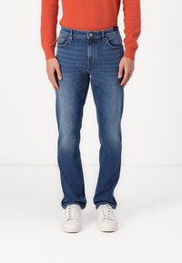 Jeans de ganga azuis com corte reto, apresentando bolsos frontais e fecho com botão. Usados com um suéter vermelho e ténis brancos.