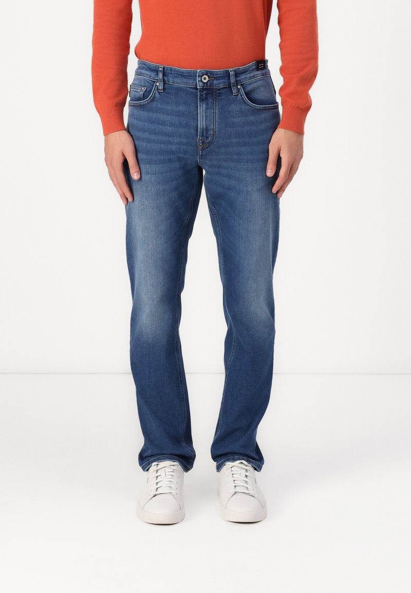Jeans de ganga azuis com corte reto, apresentando bolsos frontais e fecho com botão. Usados com um suéter vermelho e ténis brancos.