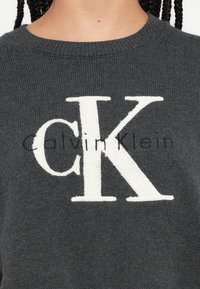 Pulover tricotat gri cu un logo alb proeminent "CK" și textul "Calvin Klein" în negru, gât rotund și material texturat.
