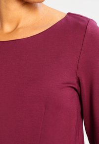 Läheltä otettu kuva henkilöstä, joka pitää yllään tiukasti istuvaa pitkähih gaalahta vaaleanpunaista paitaa, jossa on leveä, pyöreä kaula-aukko.