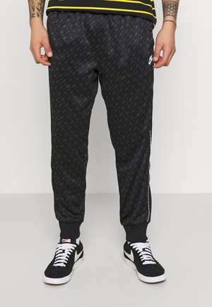 Mężczyzna w czarnych spodniach dresowych Nike z subtelnym wzorem logo, czarnych i białych butach Nike oraz czarno-żółtej koszuli w paski.