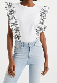 Vit t-shirt med stora svarta blomsterbroderade volanger längs ärmarna, kombinerad med ljusblå högmidjade jeans.